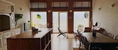 Uma sala de estar ampla com uma grande mesa de jantar de madeira, uma cozinha com armários brancos e vista para o oceano por meio de amplas portas de correr.