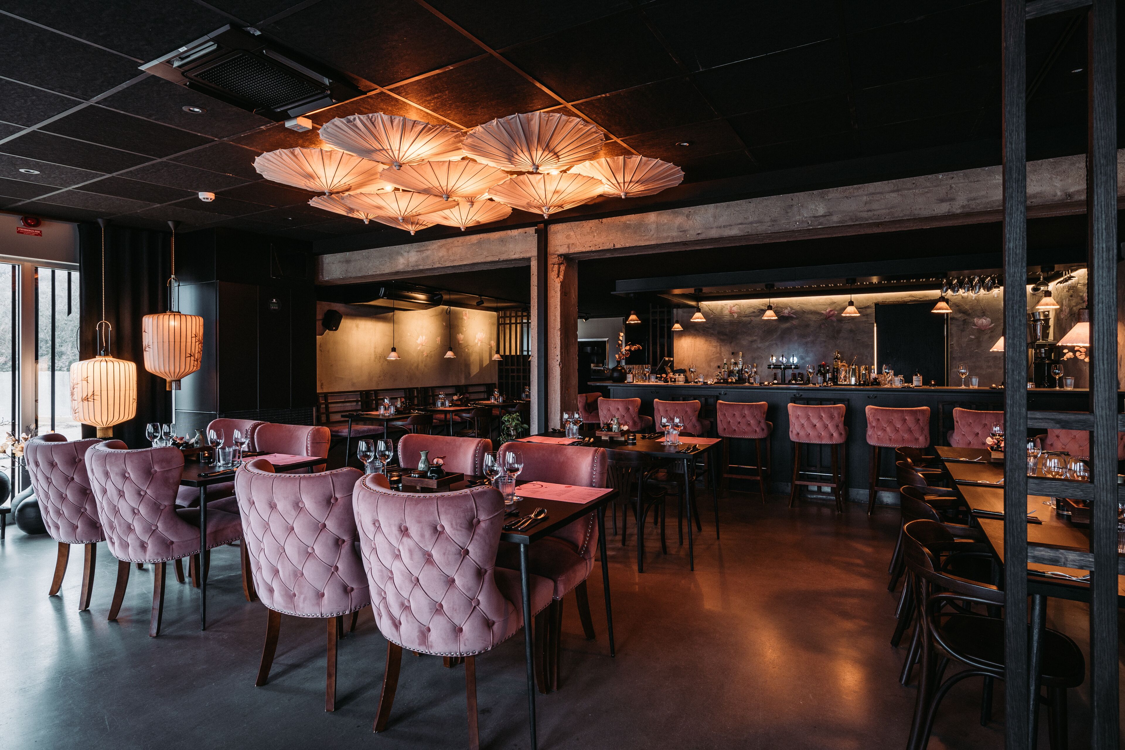 A restaurant with pink upholstered chairs, a bar area, and pendant lighting.