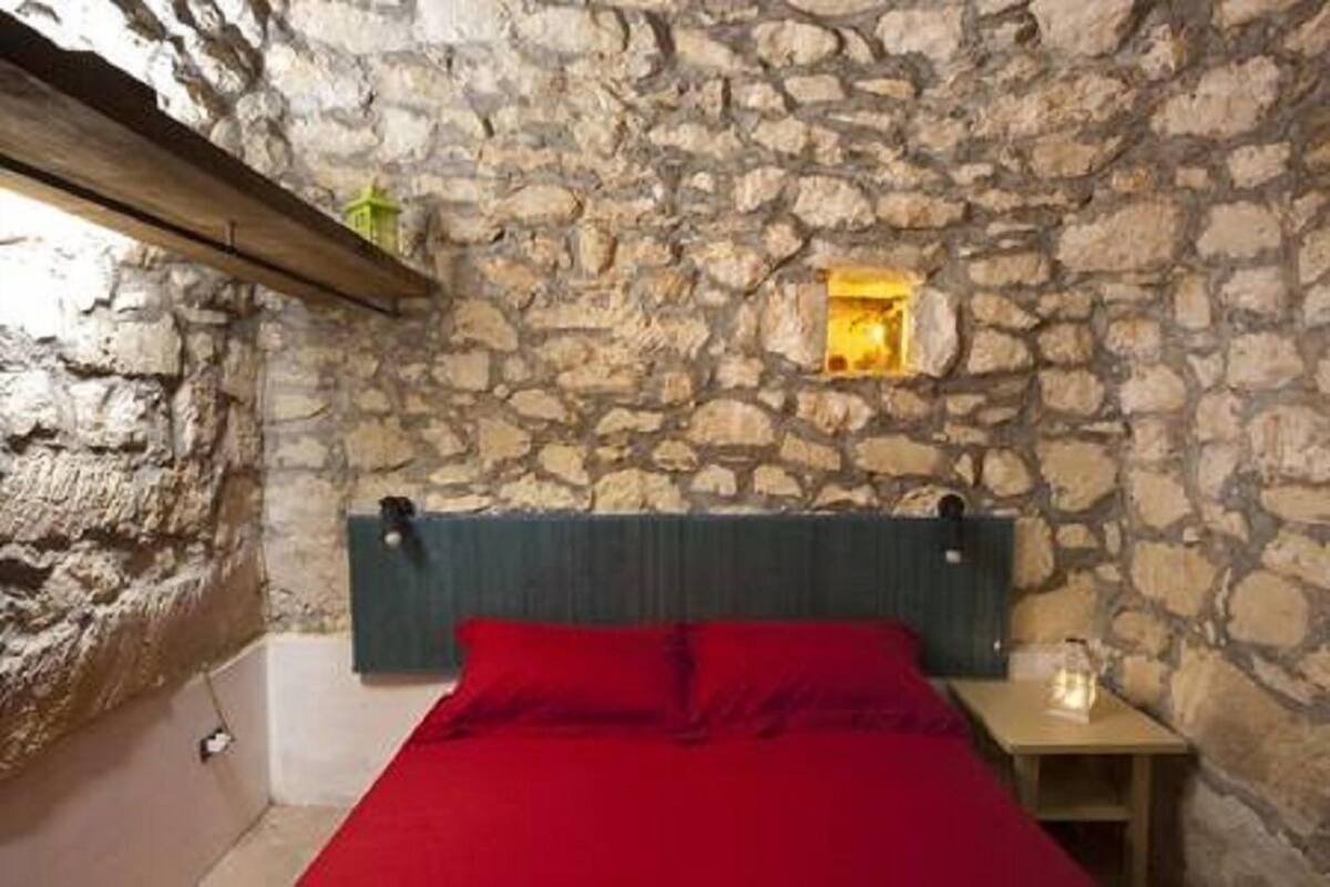 A bedroom with a stone wall, a wooden headboard, a red bedspread, and a small bedside table with a lamp.