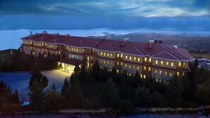 Un hotel grande de varios pisos con tejado de tejas rojas, rodeado de árboles y con vista a una masa de agua.