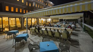 Un área de comedor al aire libre con mesas y sillas, una sección cubierta y un edificio con ventanas iluminadas al fondo.