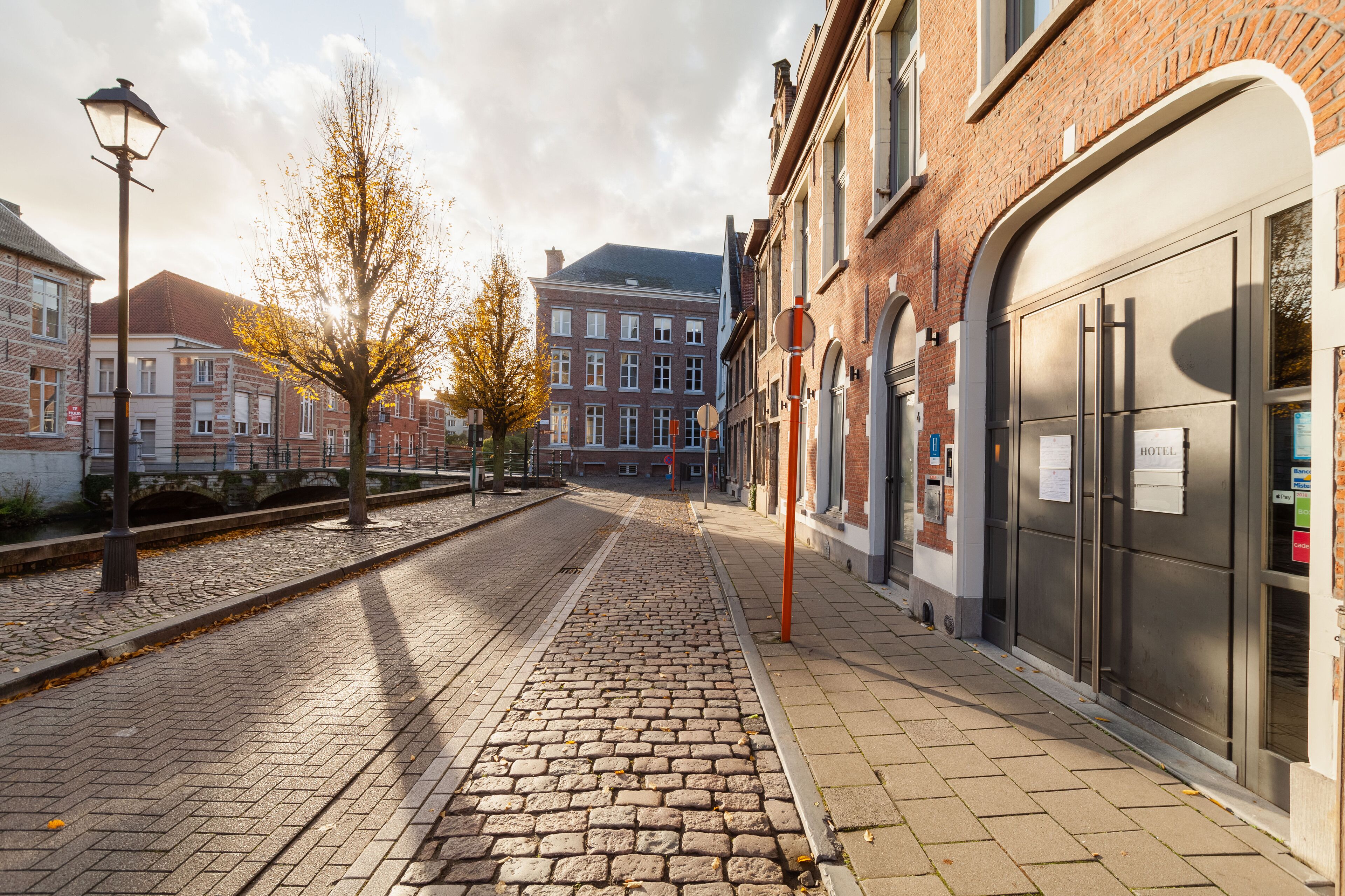 A cobblestone street with a brick building on the right, a lamppost, and a tree with autumn leaves.