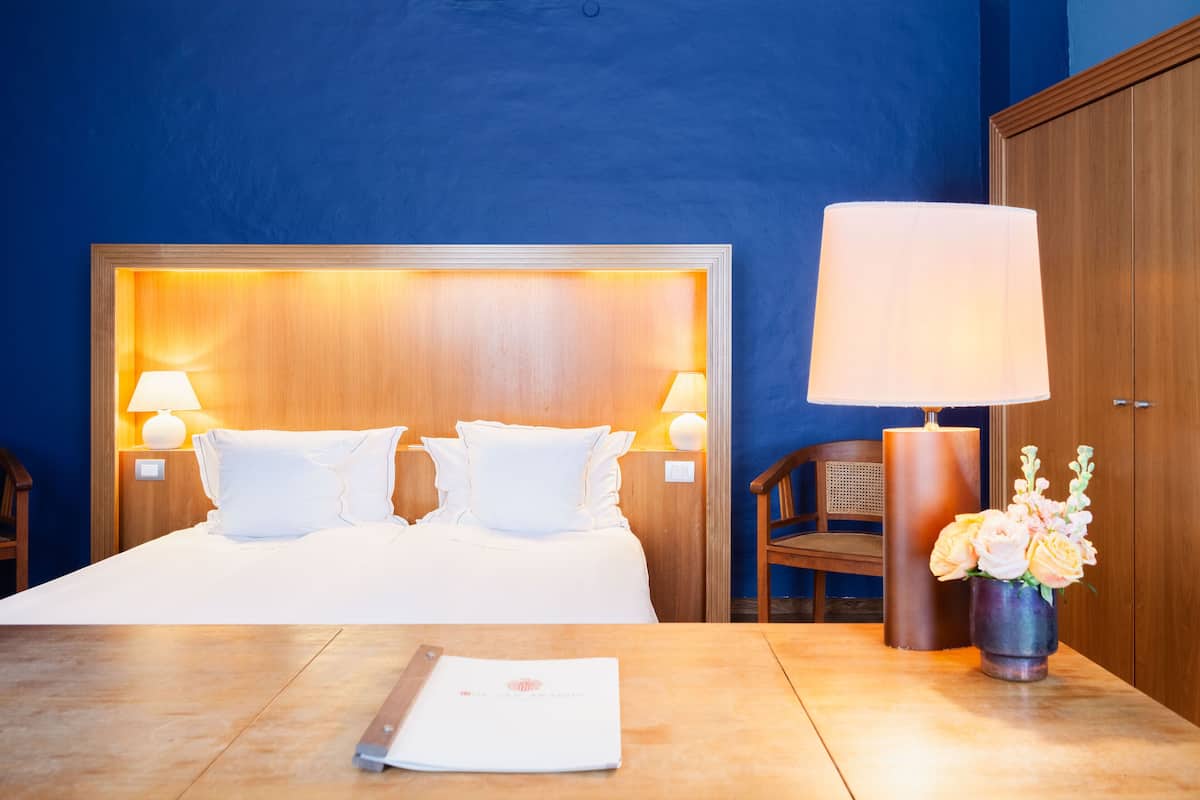 A hotel room with a large bed, two bedside lamps, a nightstand, a wooden headboard, a large candle, a vase with flowers, and a table with a menu.