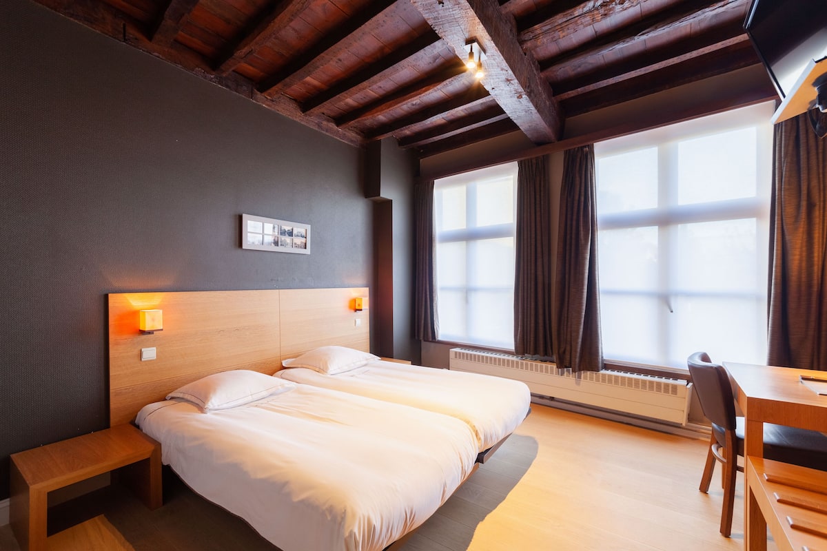 A double bed with a wooden headboard, bedside lamps, a small wooden bench, a desk with a chair, and a large window with curtains.