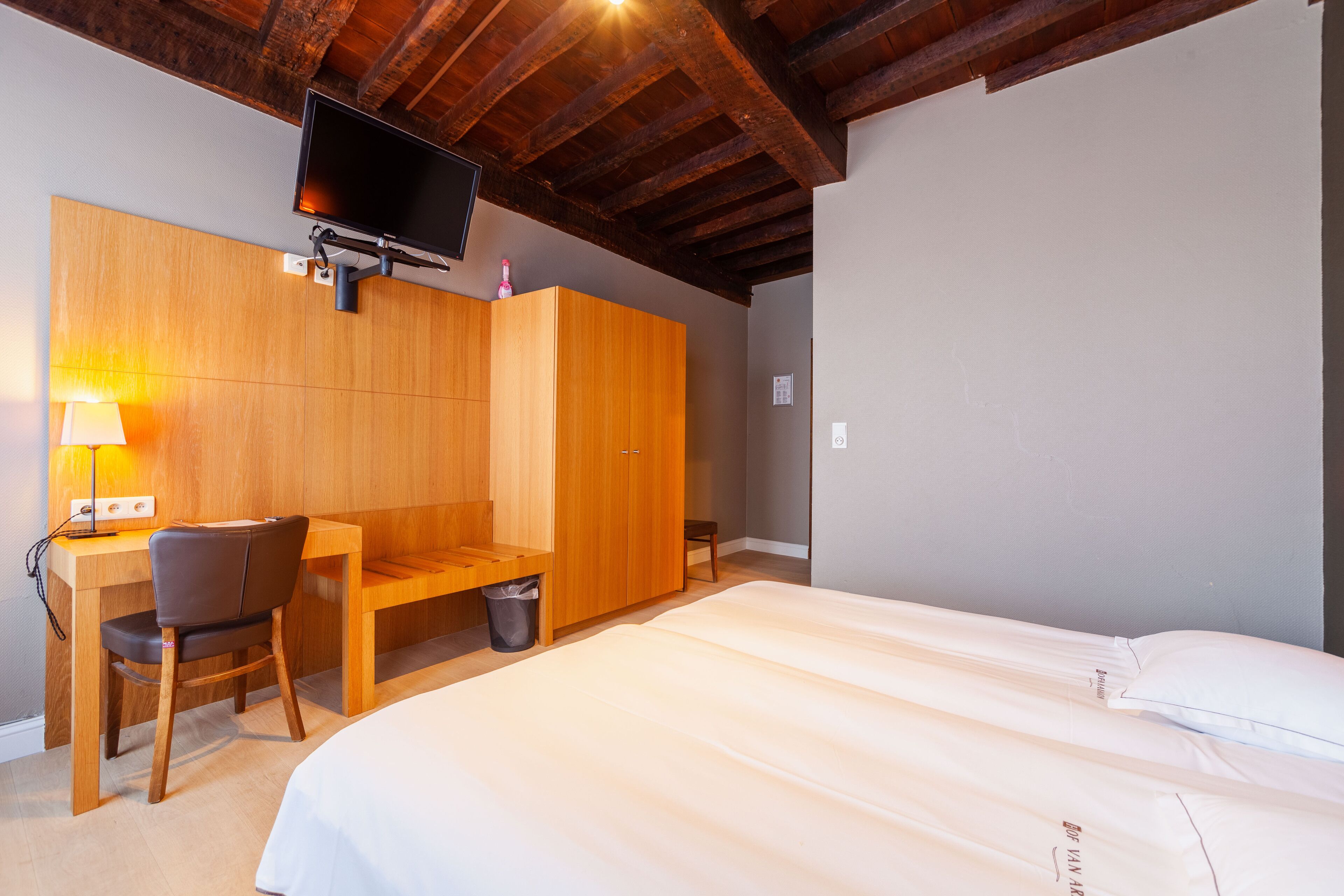 Ein Hotelzimmer mit einem Holztisch, einem Stuhl, einem Kleiderschrank, einem Bett mit weißer Bettwäsche und einem an der Wand befestigten Fernseher.