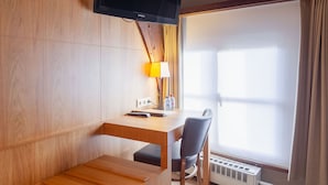 A hotel room with a wooden desk, a chair, a television mounted on the wall, a lamp, and a window with curtains.