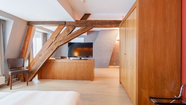 A modern hotel room with a sloped ceiling, wooden beams, a large wardrobe, a bed, a desk with a flat-screen TV, and a chair.