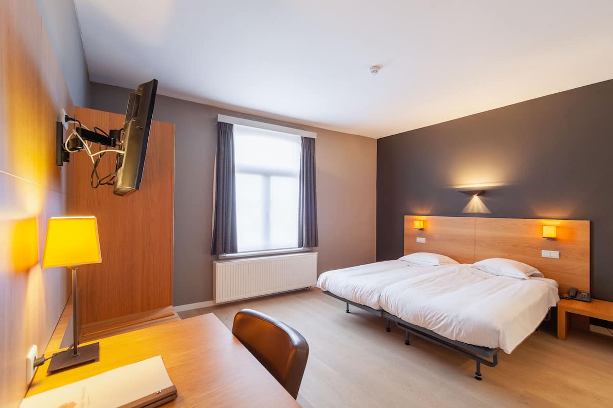 A hotel room with a bed, bedside table, lamp, desk, chair, and a TV mounted on the wall.