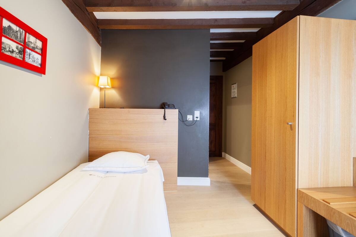 A hotel room with a single bed, a wooden wardrobe, a desk, and a wall-mounted lamp.