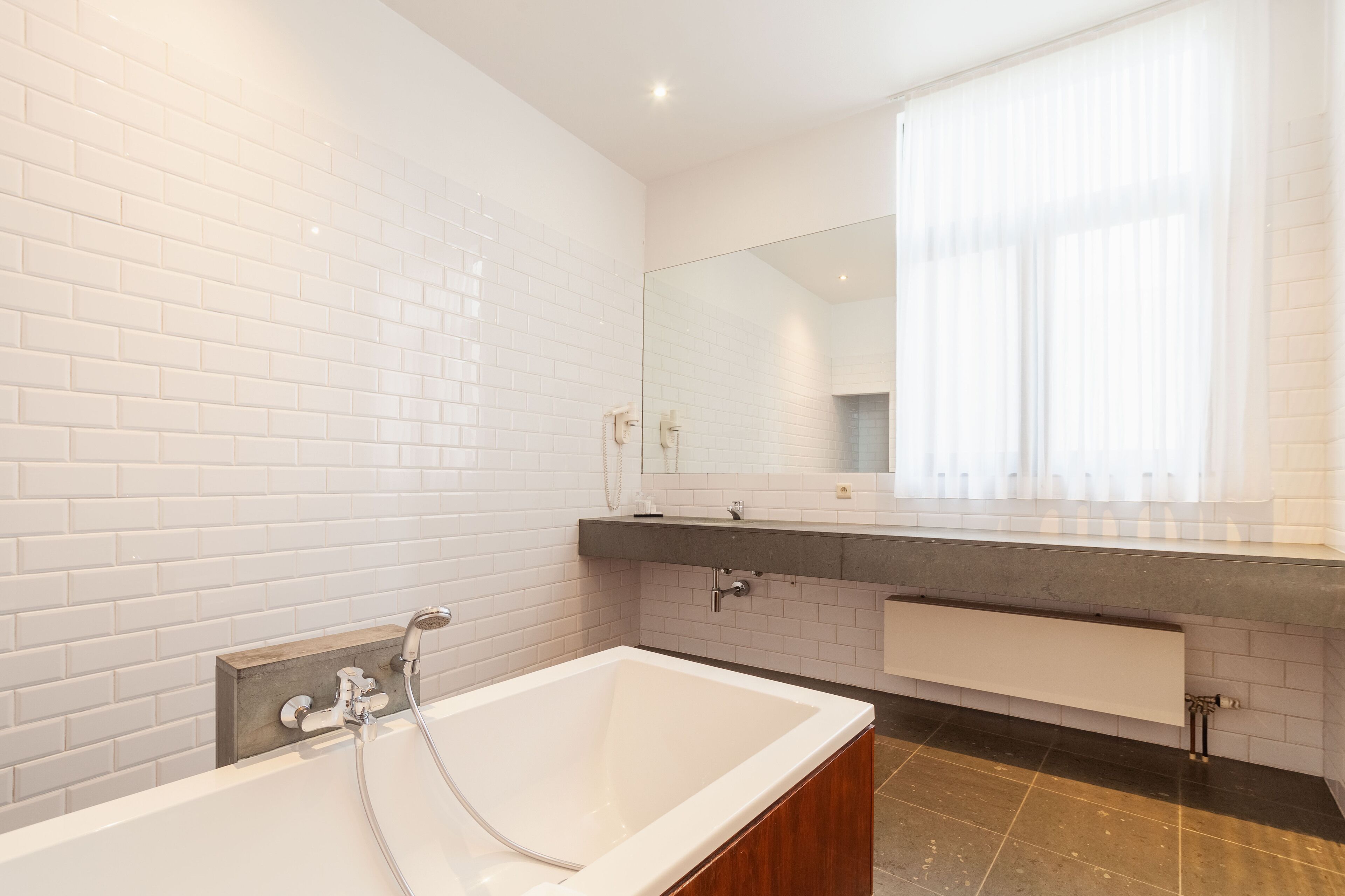 Een moderne badkamer met witte metrotegels, een groot bad, een wastafelmeubel met spiegel en een raam met dunne gordijnen.