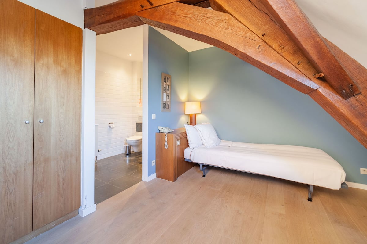 A bedroom with a wooden ceiling, a bed, a bedside table with a lamp, and a bathroom visible through an open door.