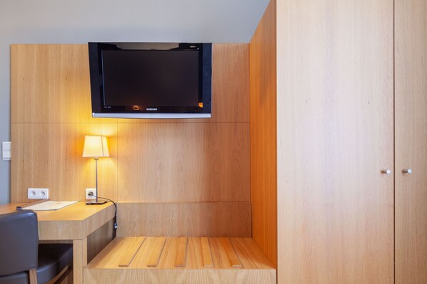 A modern hotel room with a flat-screen TV mounted above a wooden desk, a lamp, and a wooden bench.