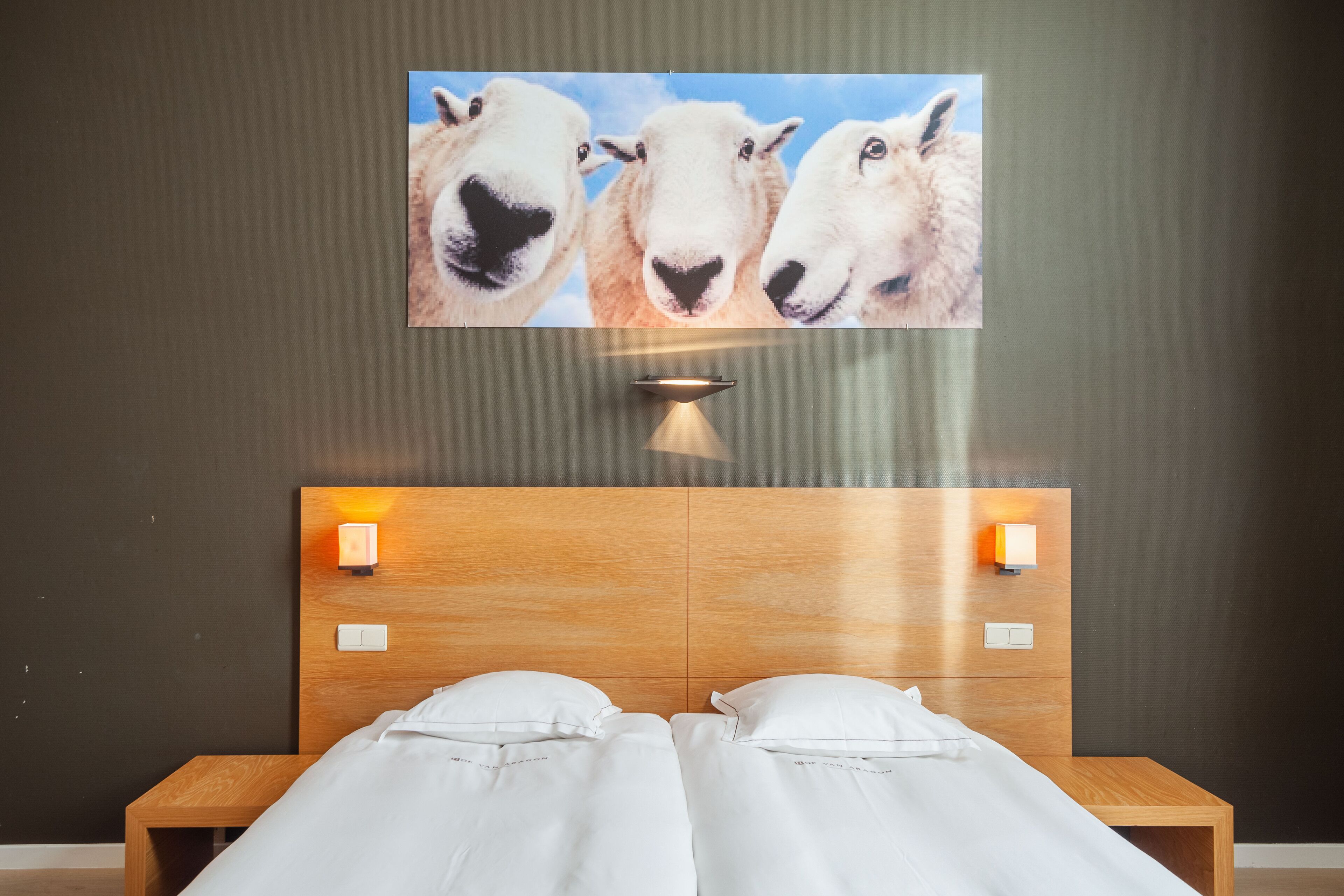 A double bed with white linens, a wooden headboard, two wall-mounted lights, and a painting of sheep above the bed.
