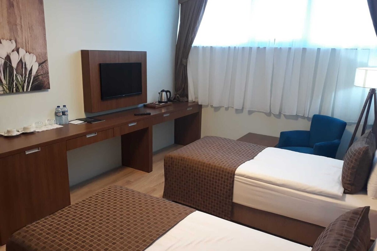 Quarto de hotel com escrivaninha de madeira, televisão, duas camas, uma cadeira azul e uma janela com cortinas.