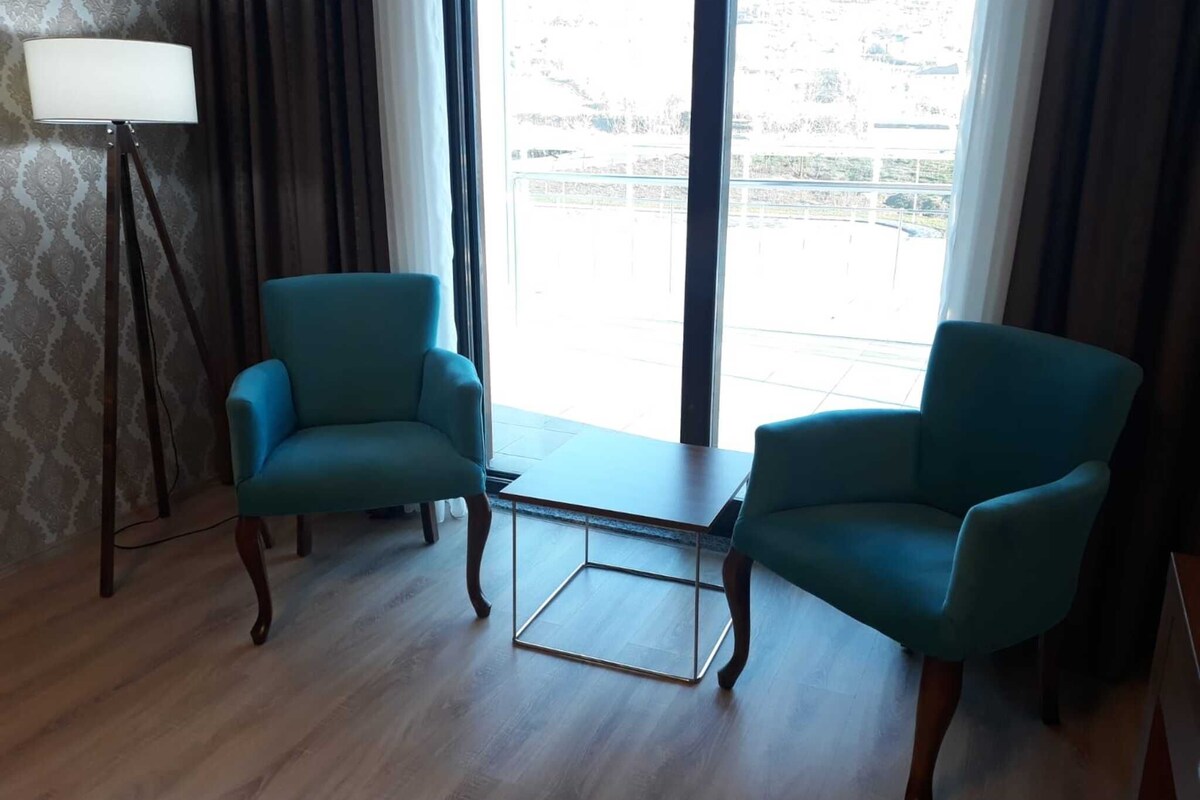 A room with two teal armchairs, a small table, a floor lamp, and a view through large windows.
