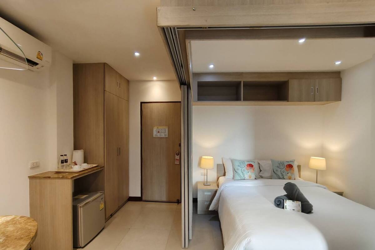A hotel room with a bed, bedside tables, a desk, a wardrobe, and an air conditioning unit.