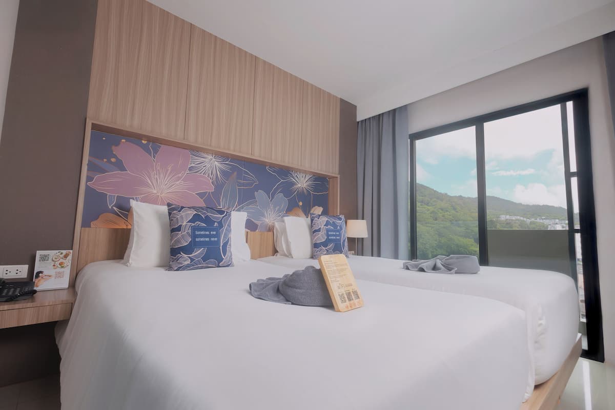 A hotel room with two beds, a large window with a city view, a wooden headboard, and a floral artwork above the beds.