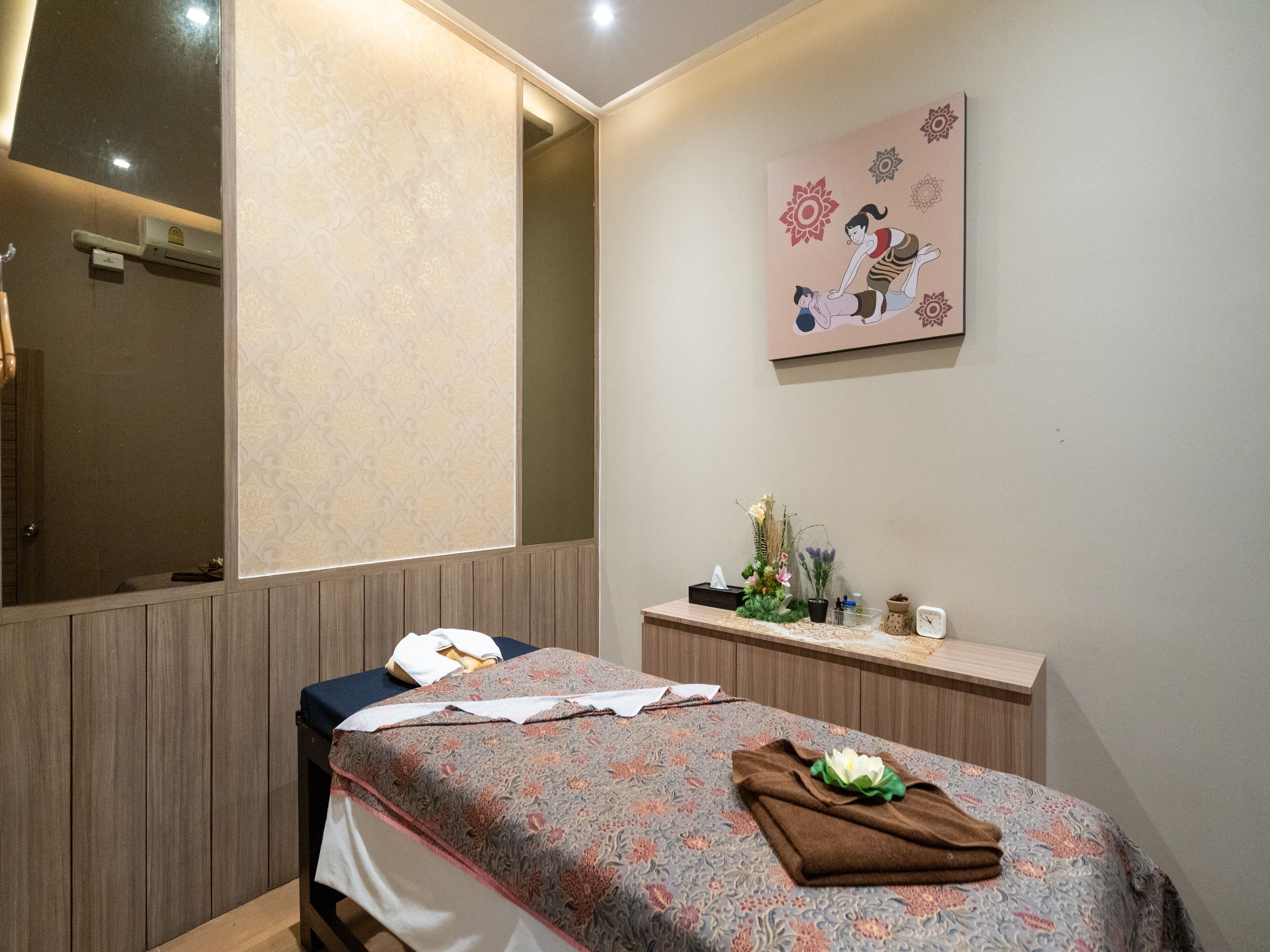 A room with a bed, a mirror, a wall with a floral painting, and a small table with a vase of flowers.