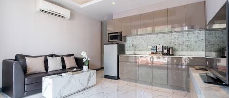 Una sala moderna con una mesa de centro de mármol, un sofá con cojines, y una zona de cocina con microondas, lavamanos y salpicadero de azulejos.