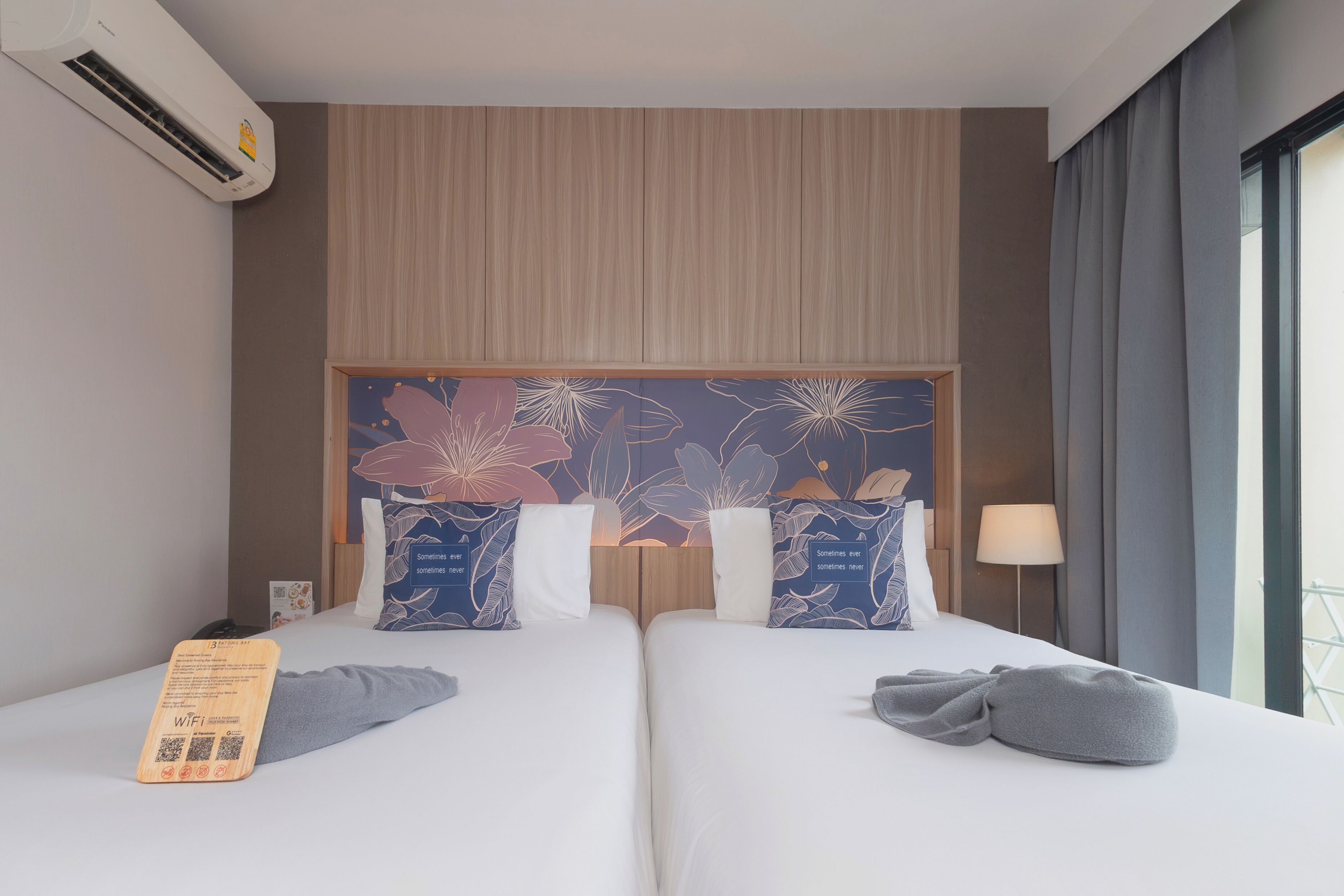 A hotel room with two beds, a wooden headboard, a floral wall mural, a window with curtains, and a bedside lamp.