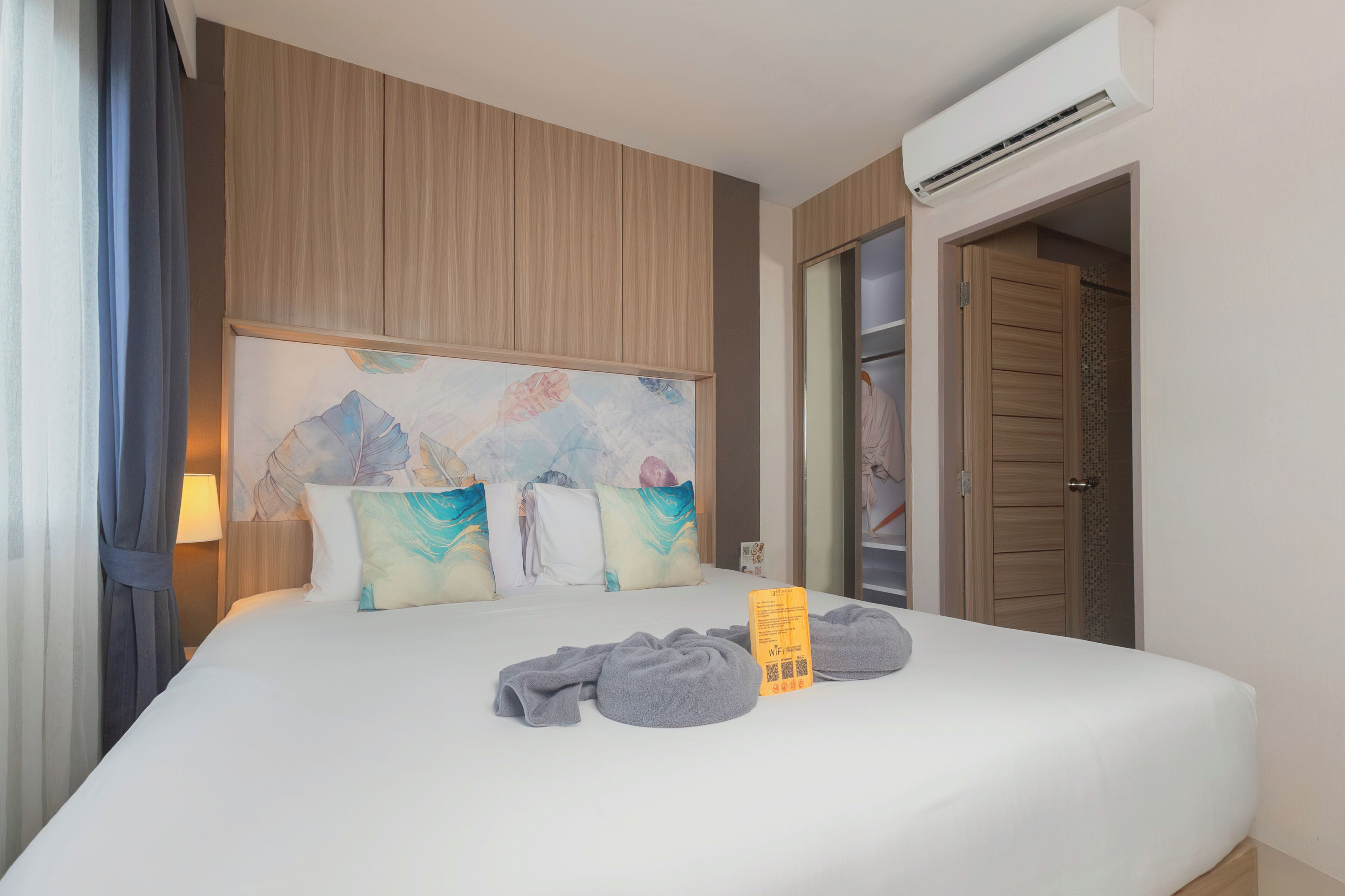 A hotel room with a large bed, a headboard with artwork, a wardrobe, and an air conditioning unit.