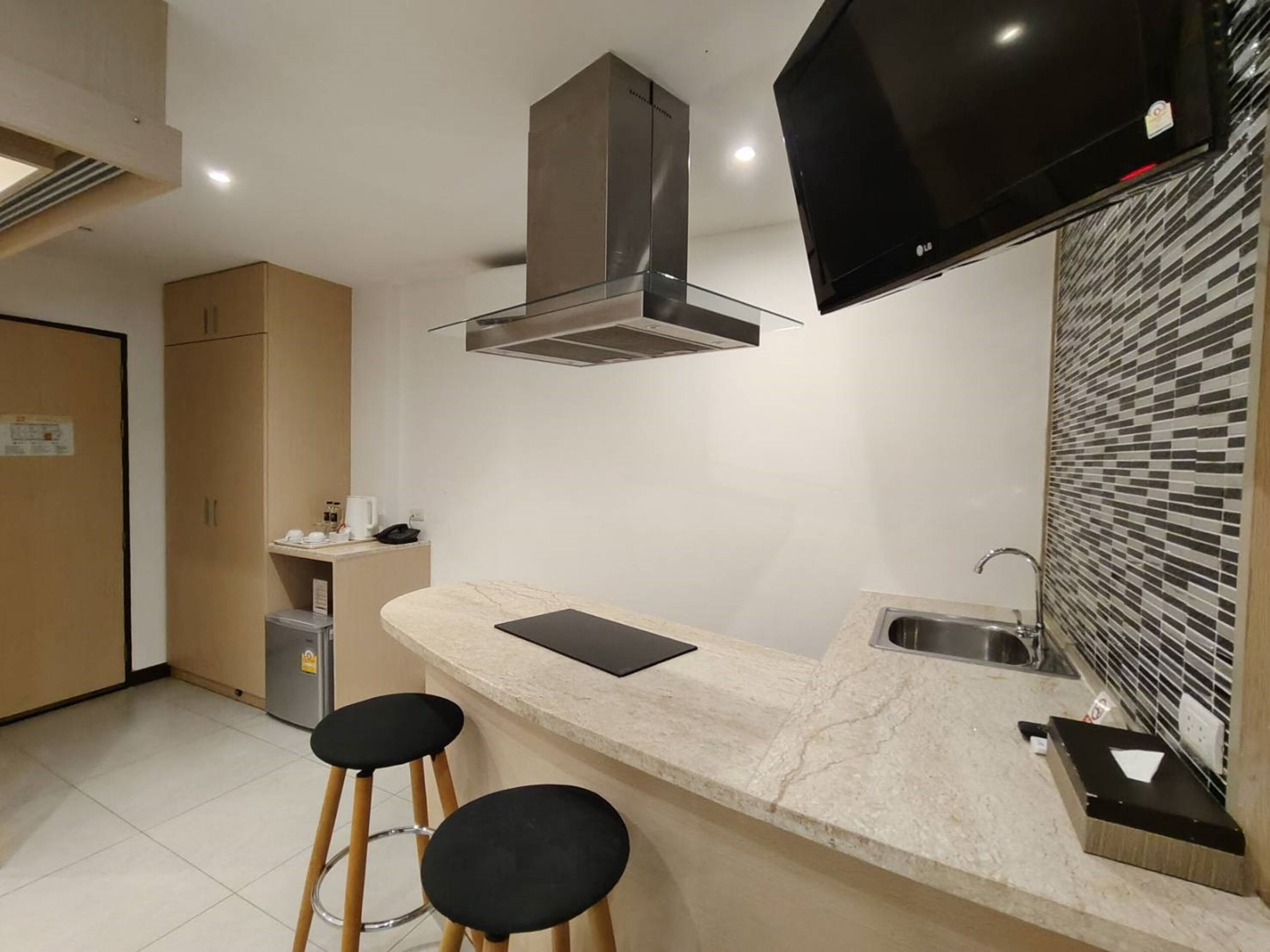 A modern kitchen with a marble countertop, two black stools, a stainless steel sink, and a flat-screen TV mounted above the counter.