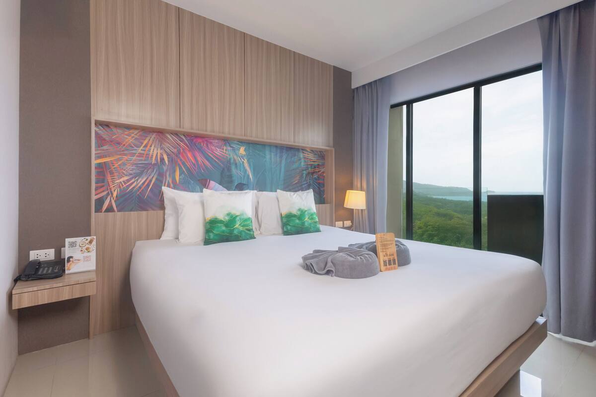 A hotel room with a large bed, a nightstand with a phone, a painting on the wall, and a view of the outdoors.