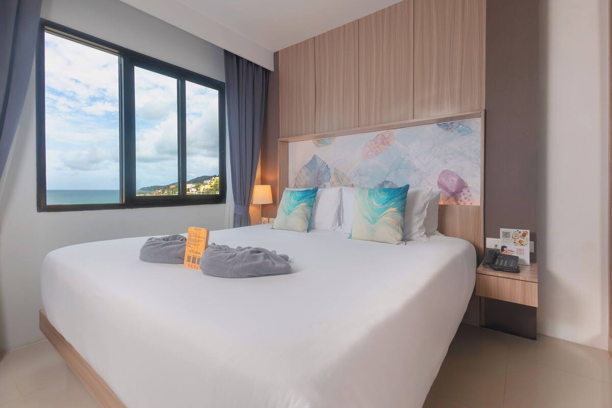 A modern hotel room with a large bed, a nightstand, a lamp, and a view of the sea.