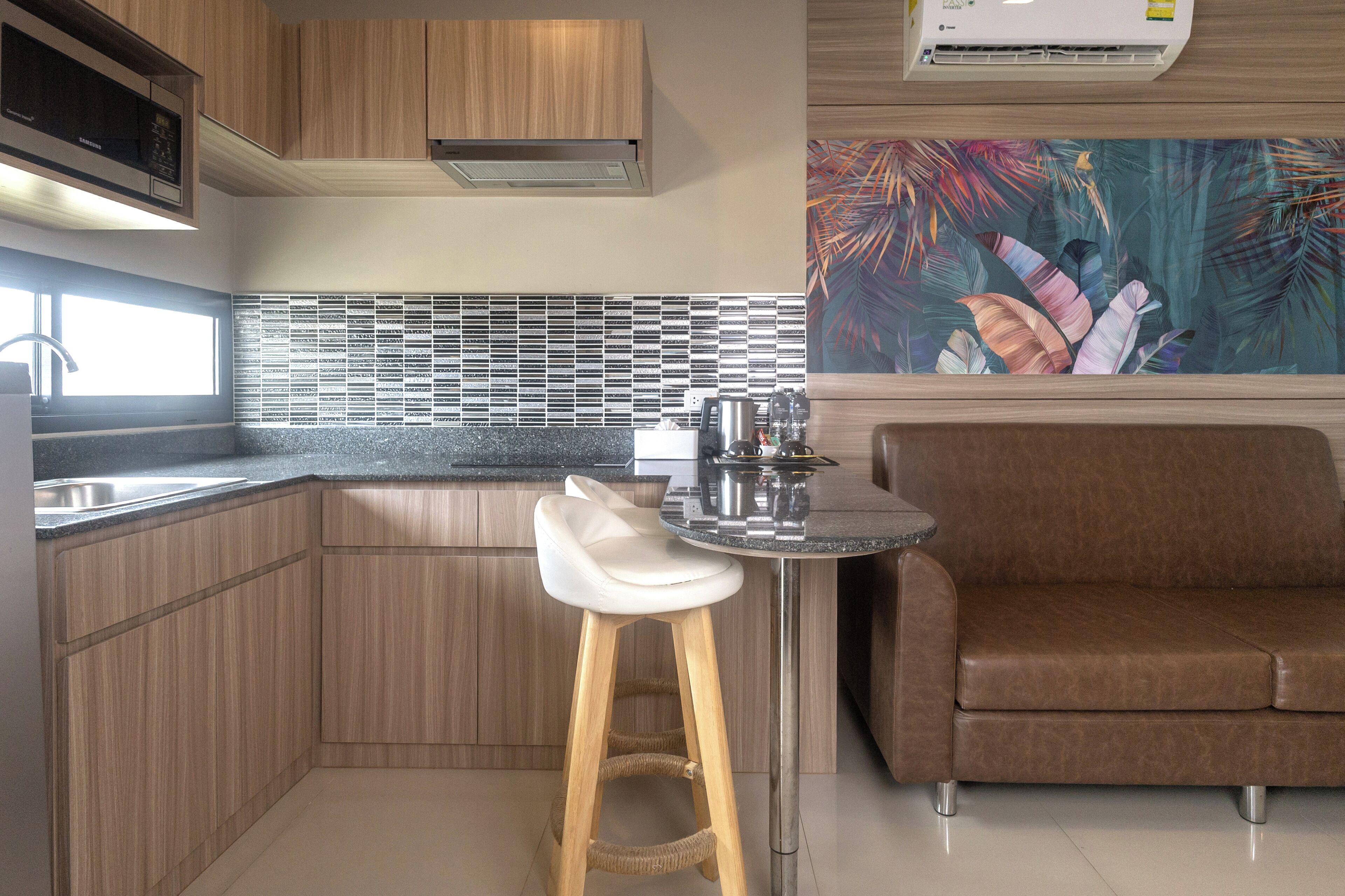 Une cuisine avec des meubles en bois, un évier en acier inoxydable, un tabouret de bar blanc, un canapé en cuir marron et un mur décoratif orné d’un motif de feuilles tropicales.
