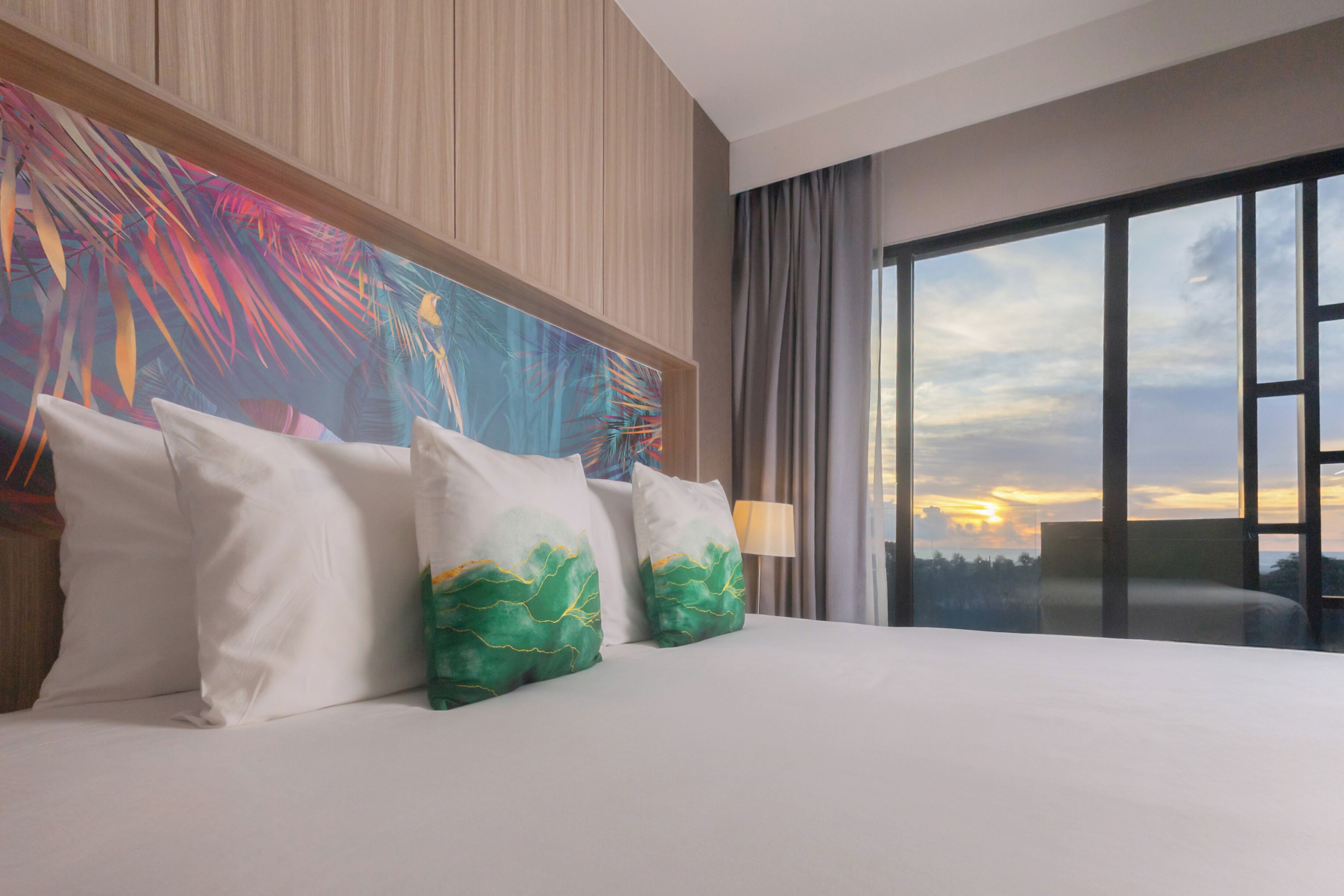 A hotel room with a large bed, a colorful abstract painting on the wall, and a view of the sunset through a large window.