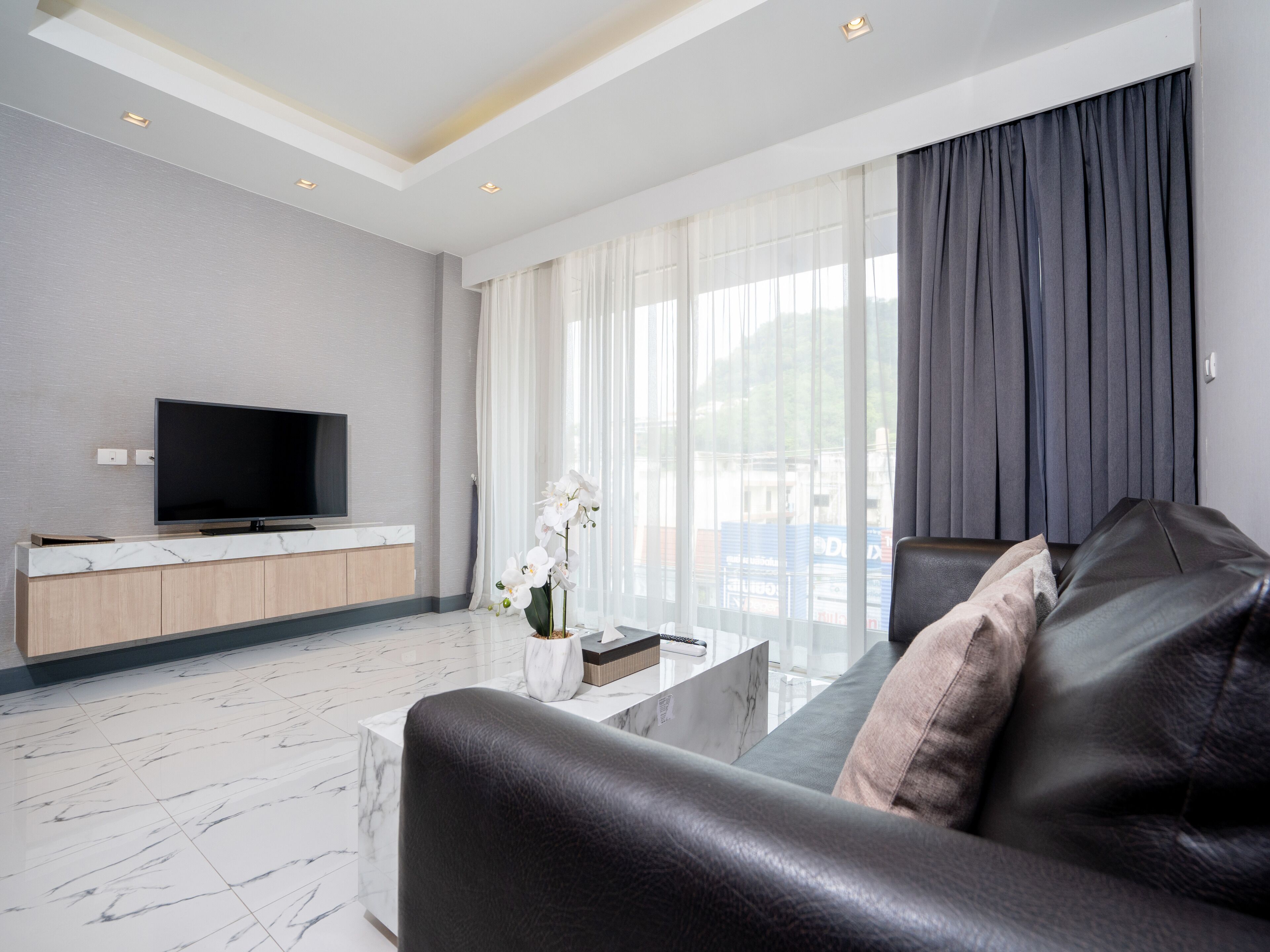 Una sala de estar moderna con un televisor de pantalla plana, una mesa de centro de mármol, un sofá de cuero y un amplio ventanal con cortinas translúcidas.