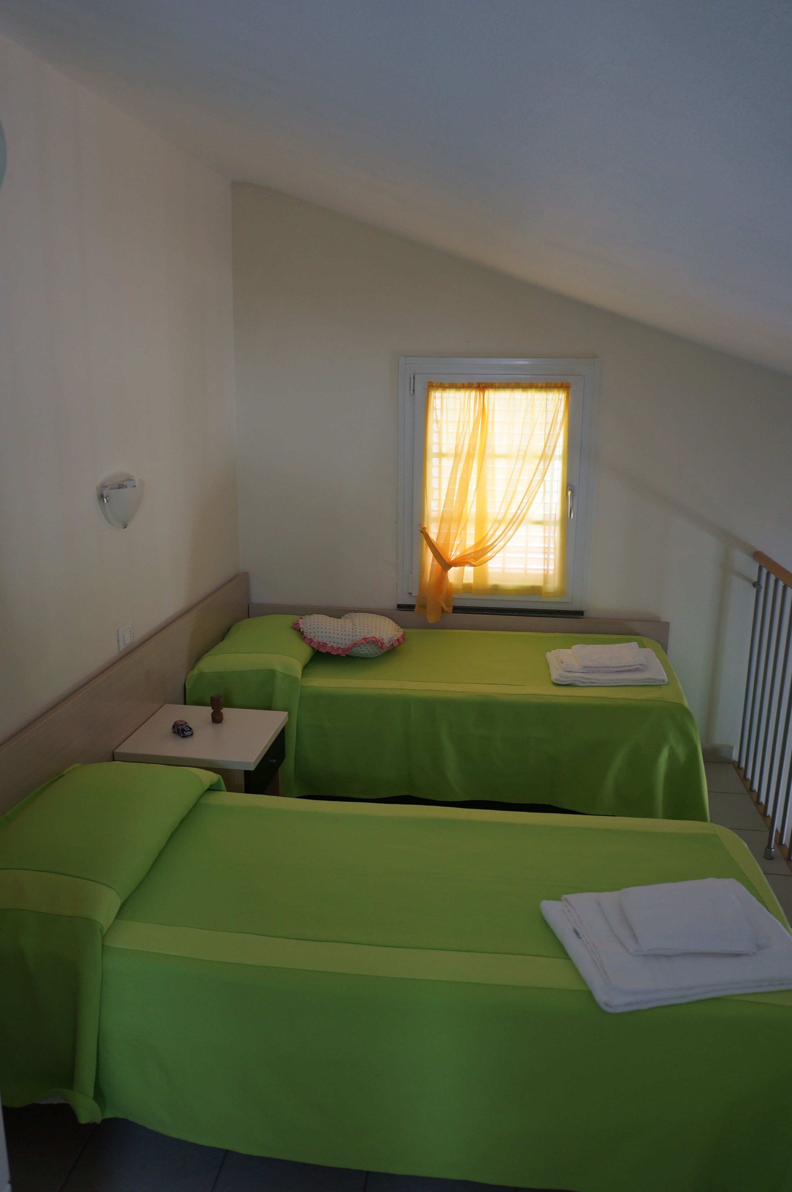 A room with two single beds, a small table, a window with curtains, and a wall-mounted light fixture.