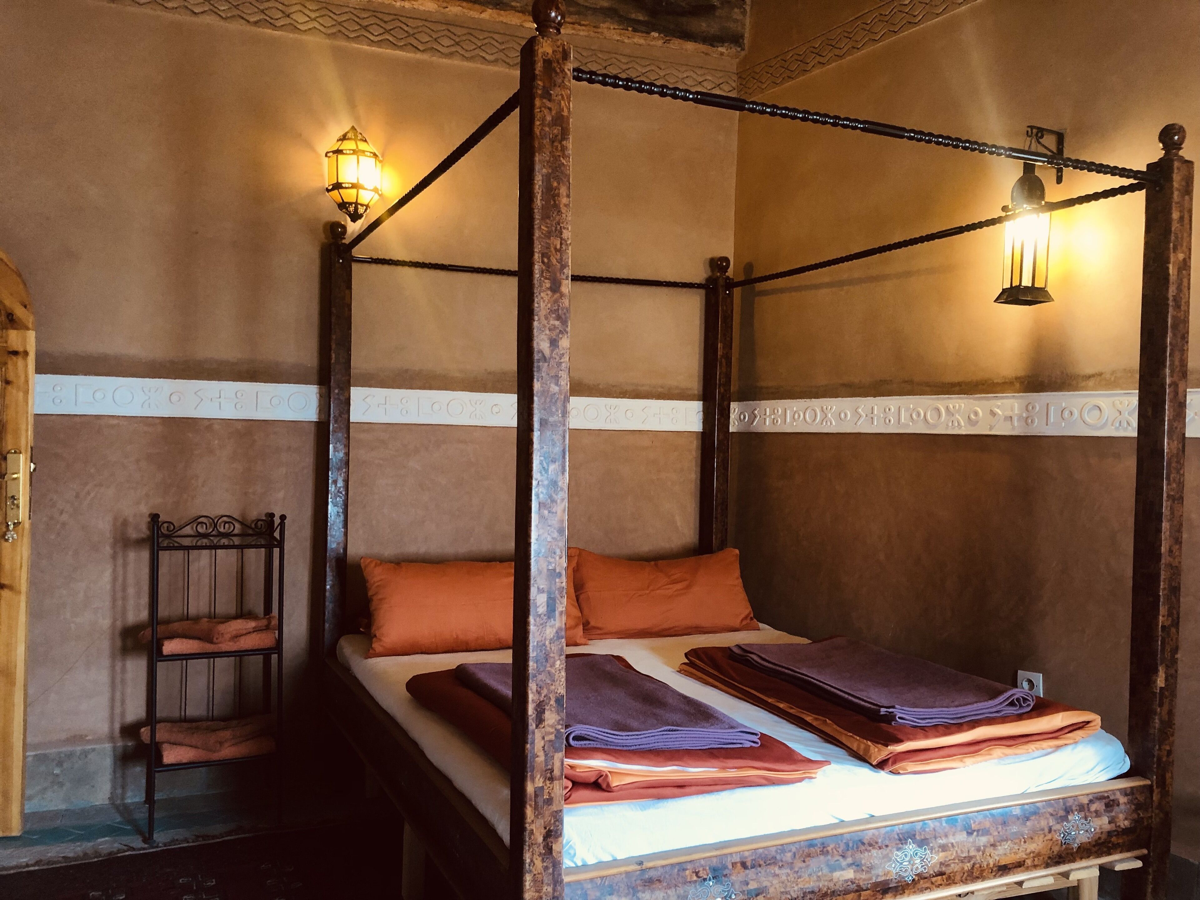 Un lit à baldaquin avec une structure en bois, une tête de lit en métal et une petite étagère où sont pliés des serviettes. Il y a deux lampes fixées au mur et un panneau décoratif.