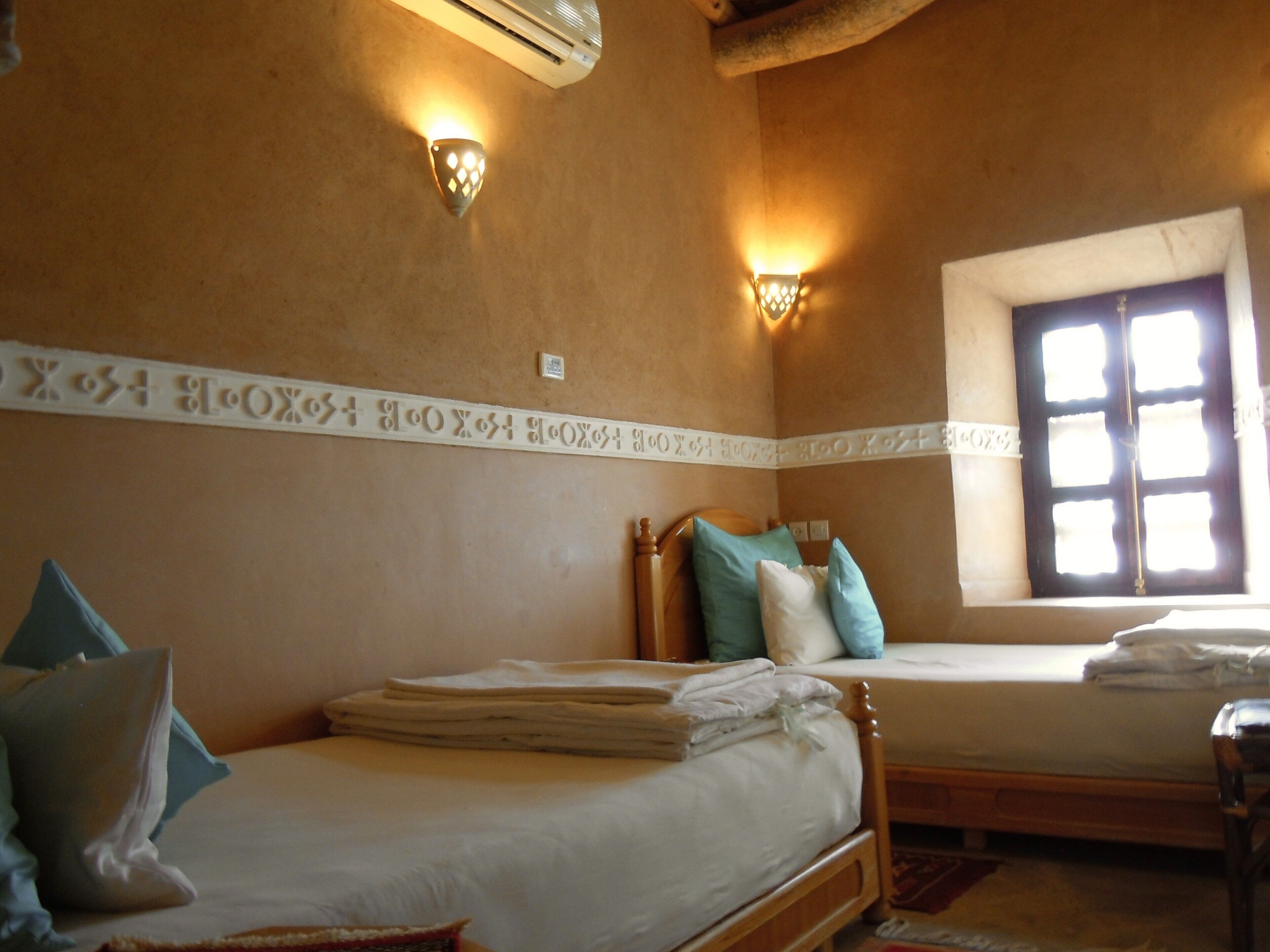 A room with two beds, a window with blinds, and wall-mounted lights.