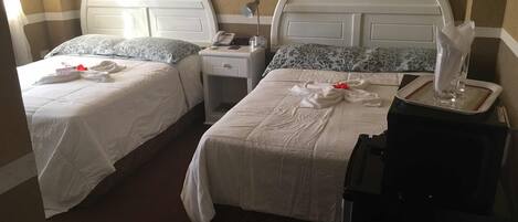 Habitación de hotel con dos camas, cada una con una colcha blanca y cabecero con un logotipo. Se observa una mesita de noche con una lámpara, un teléfono y una caja de pañuelos. En primer plano, una mesita con un vaso de agua y una caja de pañuelos.