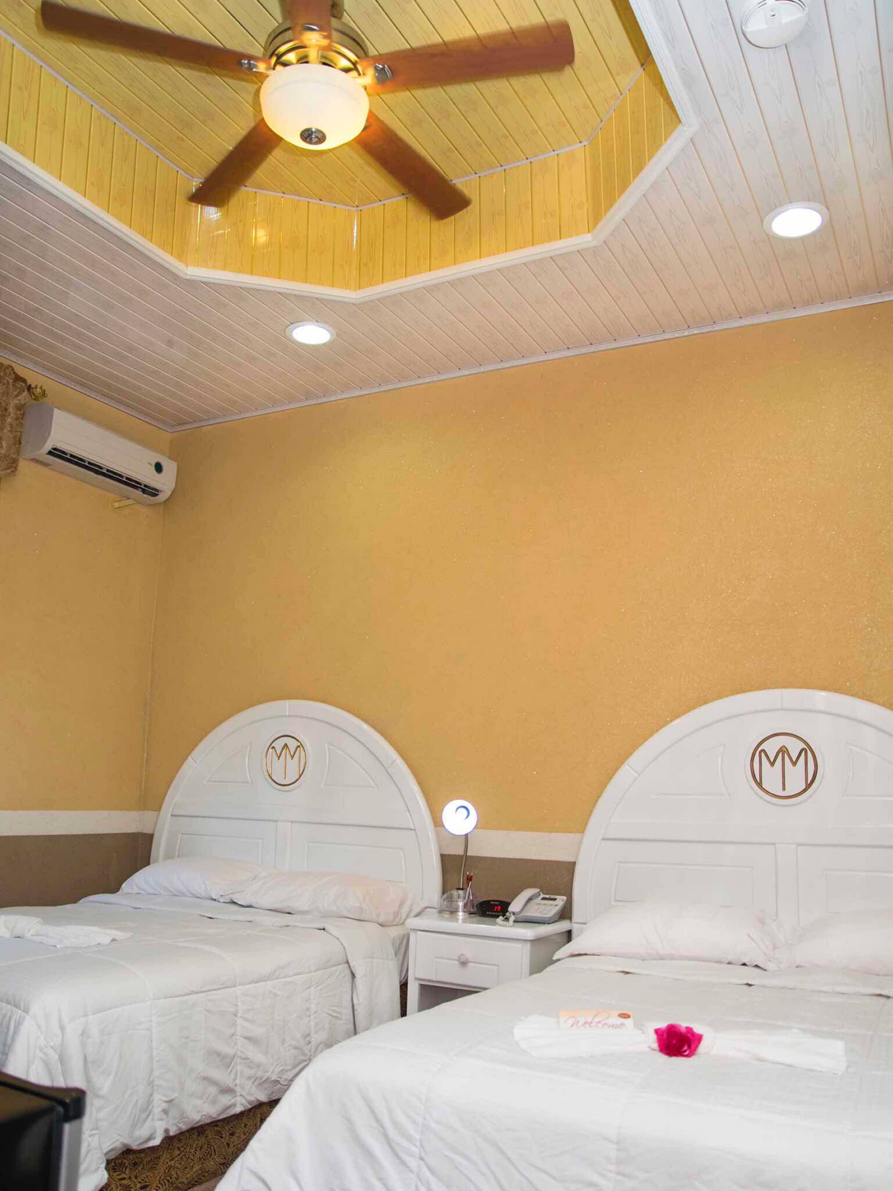 Ein Hotelzimmer mit zwei Betten, einem Deckenventilator, einer Klimaanlage und einem Nachttisch mit Telefon und Lampe.