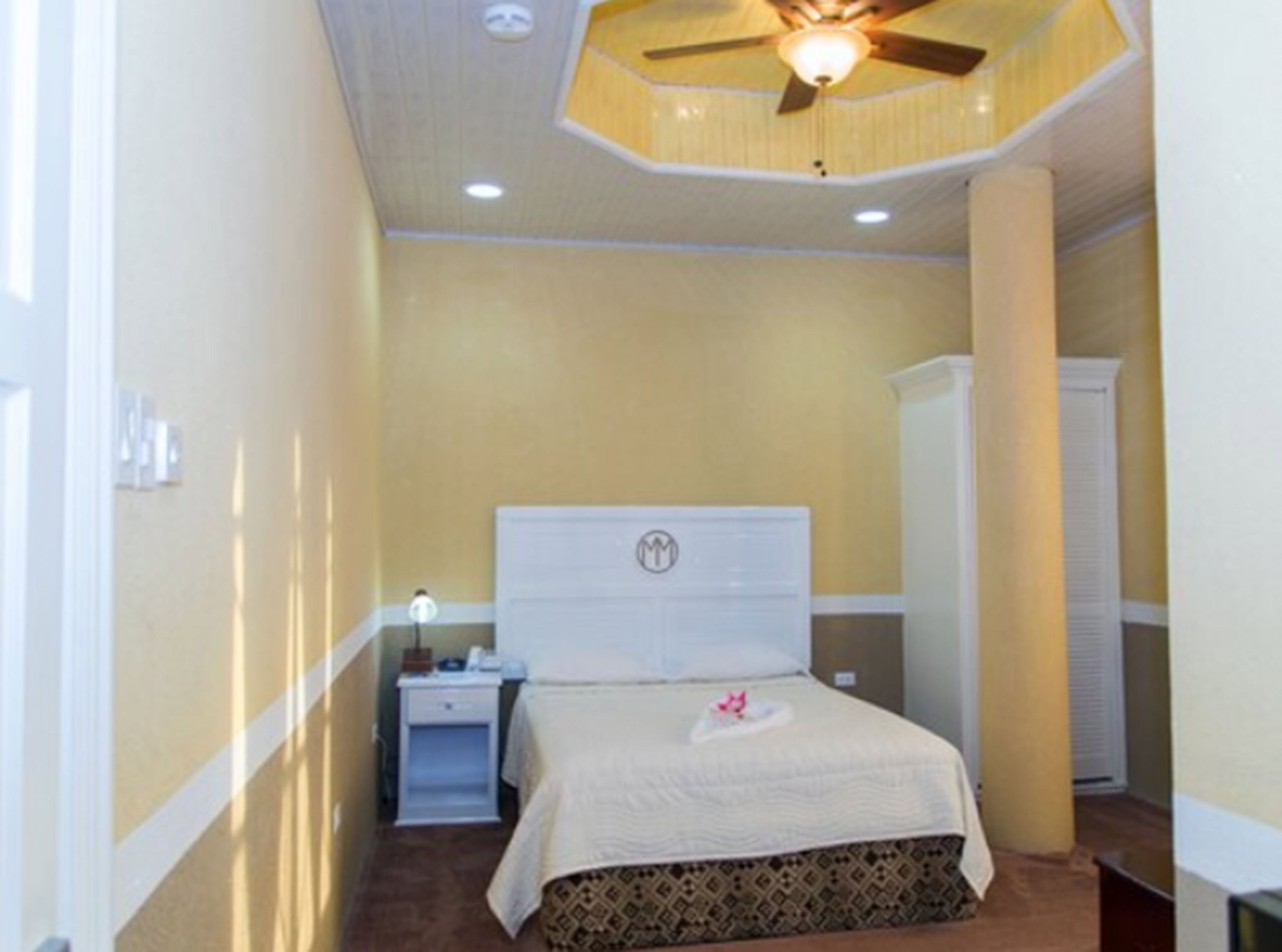 Habitación de hotel estrecha con una cama individual, una mesita de noche, un ventilador de techo y un armario blanco.