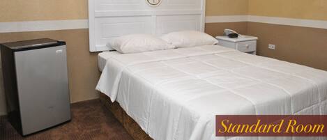 A standard room with a bed, white bedding, a nightstand, a telephone, and a small refrigerator.
