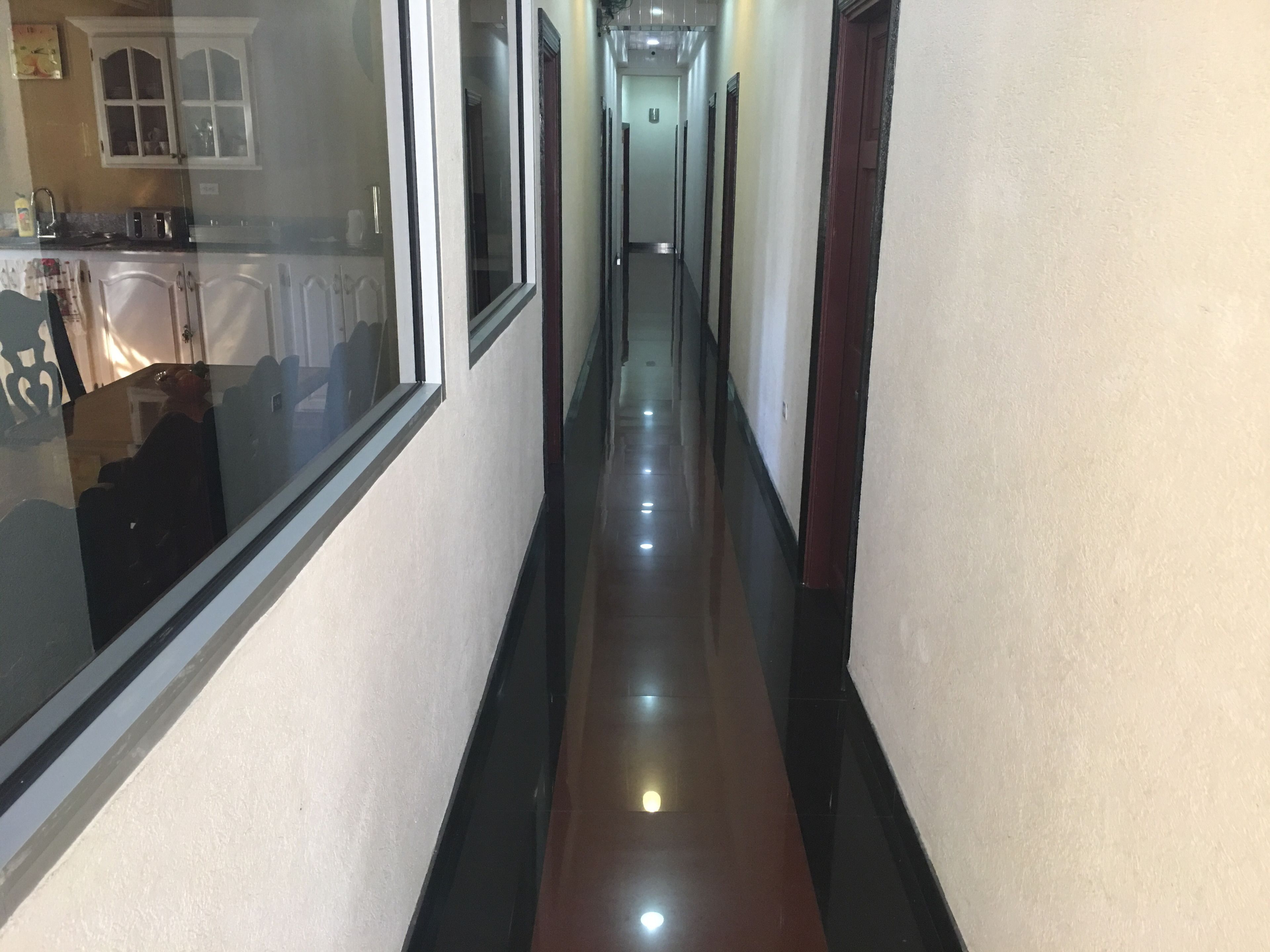 A narrow, well-lit hallway with a reflective floor, leading to a kitchen area with cabinets and a window.