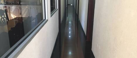 A narrow, well-lit hallway with a reflective floor, leading to a kitchen area with cabinets and a window.