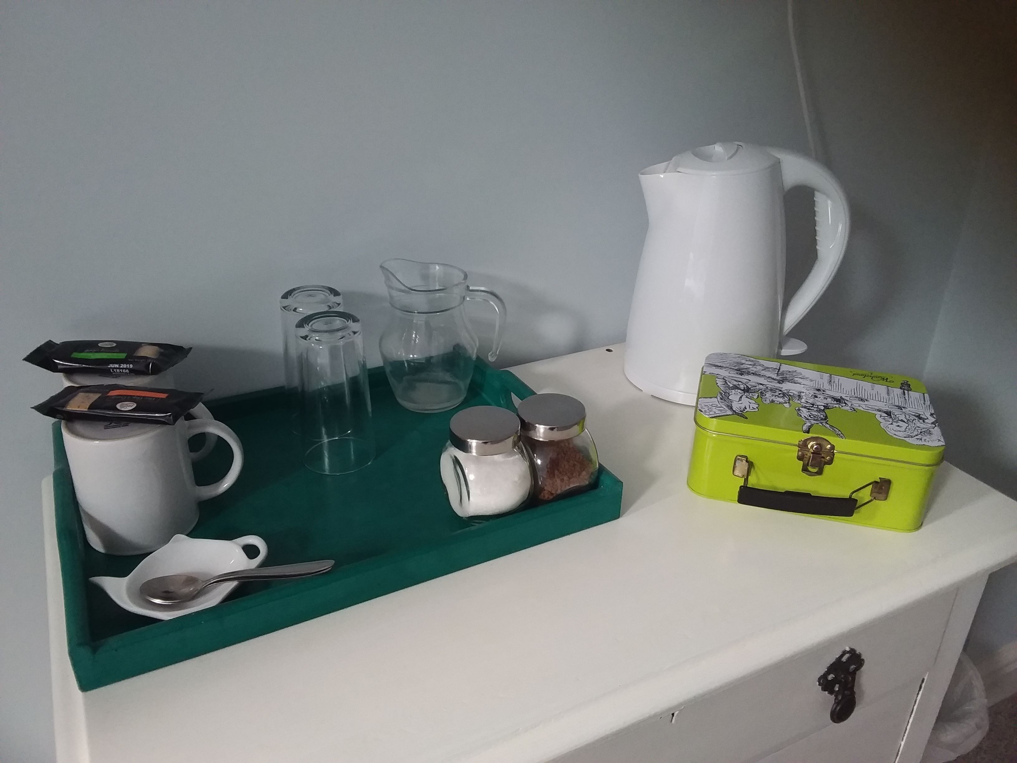 Una mesa blanca con una bandeja verde que sostiene una taza blanca, dos vasos transparentes, una jarra blanca, una caja verde y un recipiente blanco con tapa.