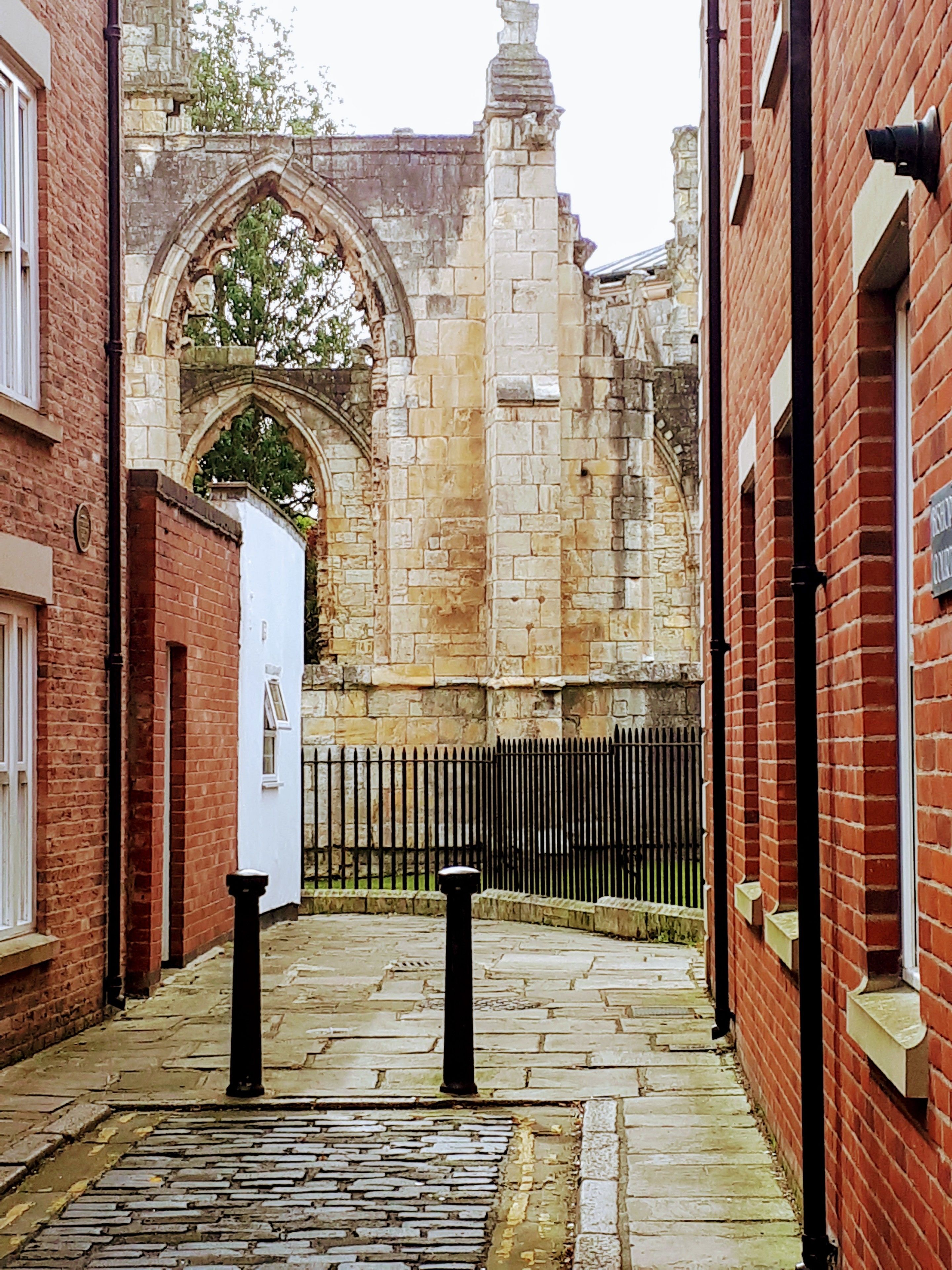A narrow alleyway with cobblestone pavement, a black metal fence, and a historic stone building with an arched entrance.