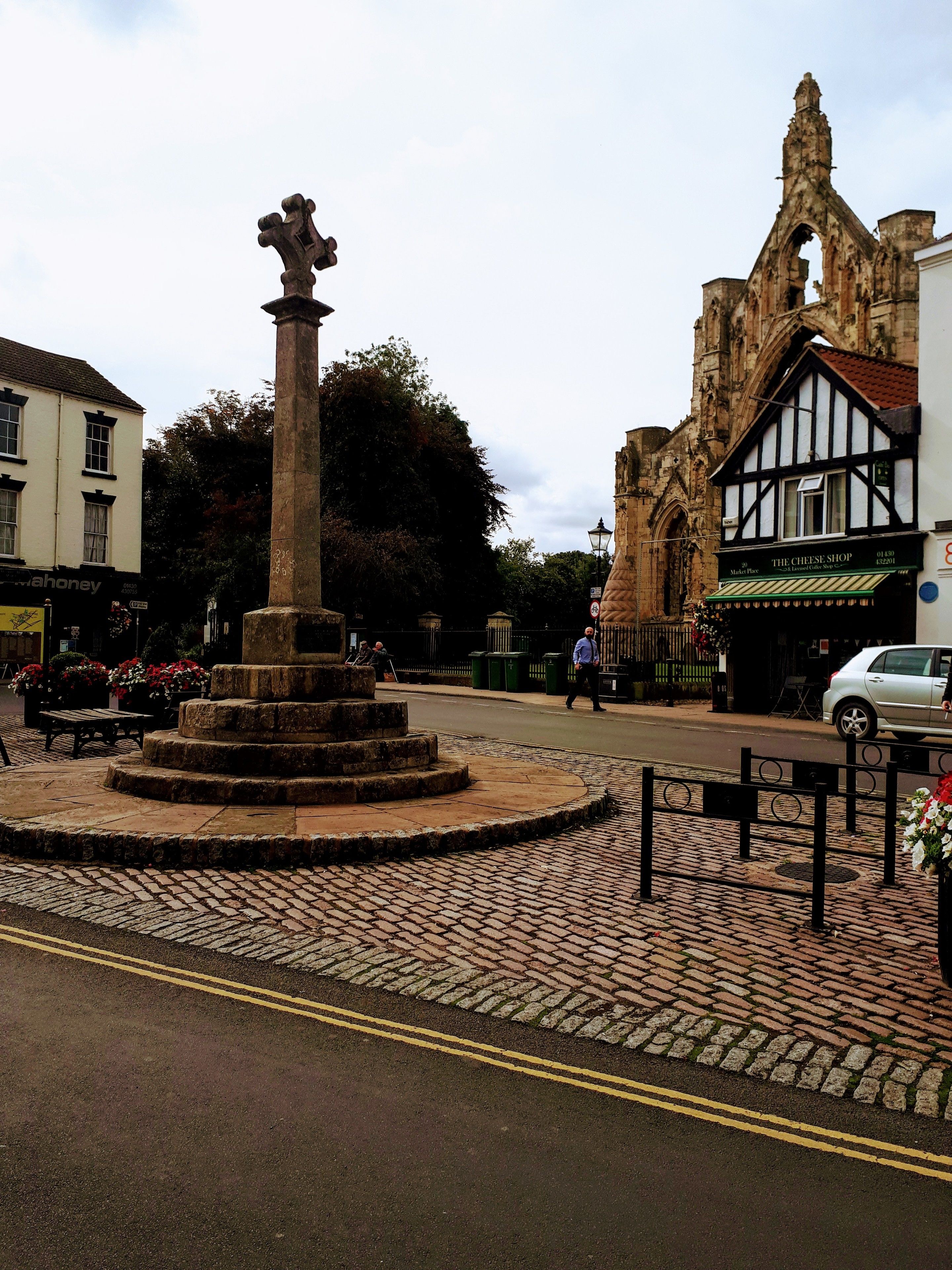 A historic stone monument in a town square with cobblestone pavement, surrounded by traditional buildings and a church with Gothic architecture.