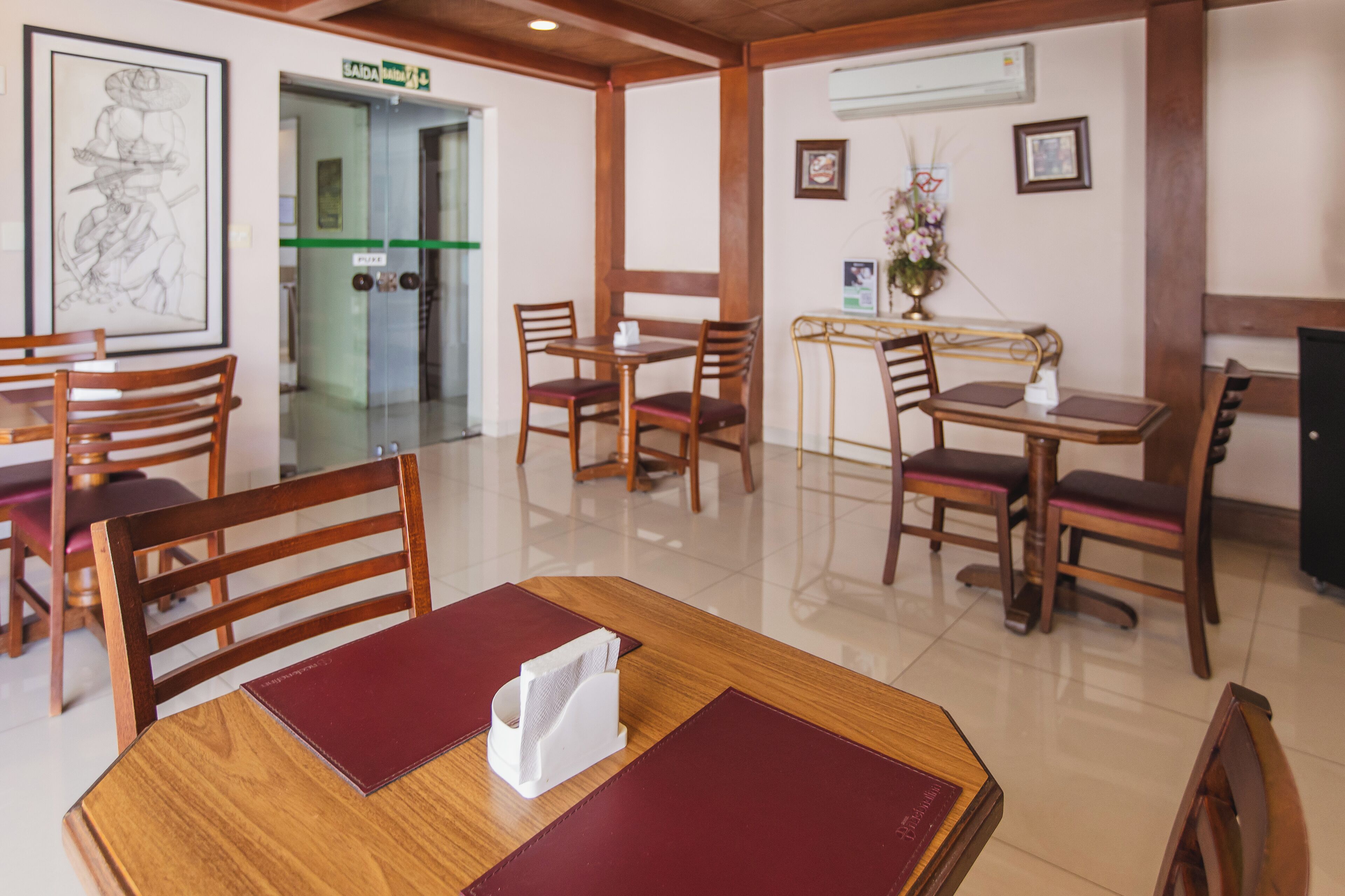 Un comedor de hotel con mesas y sillas de madera, una obra de arte enmarcada en la pared y un letrero de salida verde.