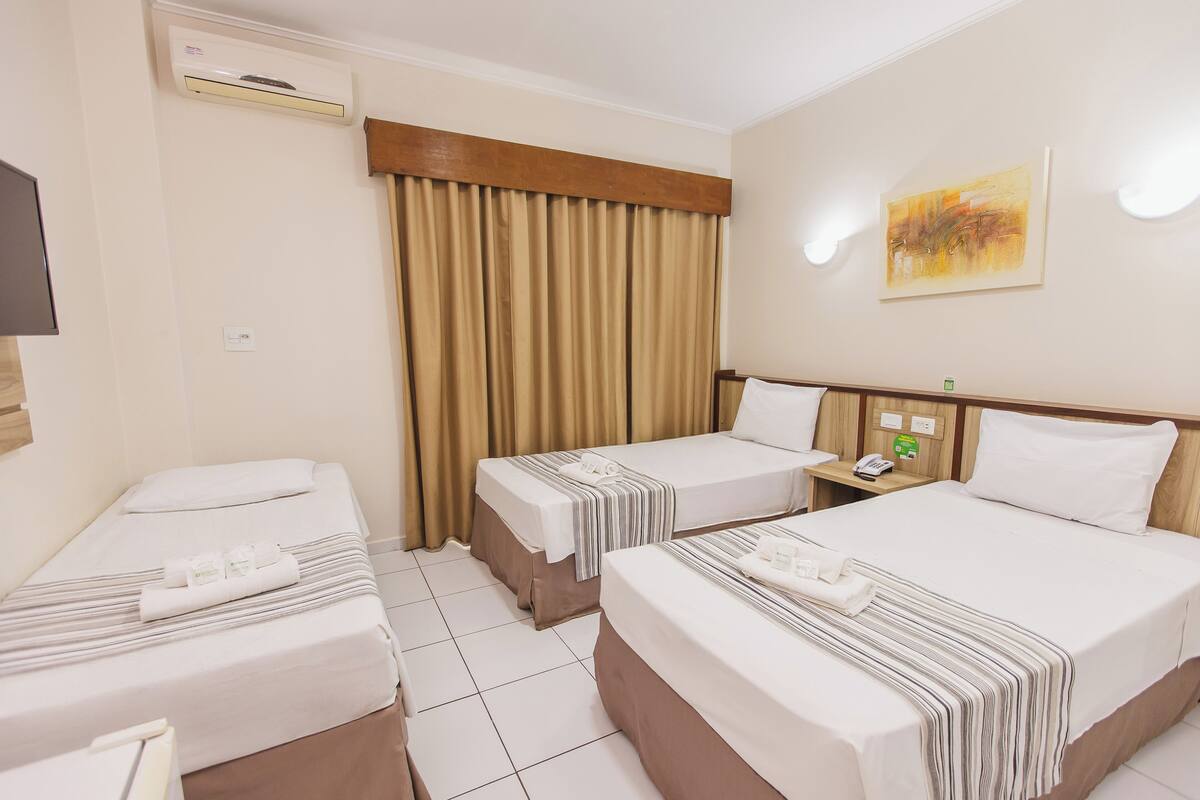 Ein Hotelzimmer mit zwei Einzelbetten, einem großen Fenster mit Vorhängen, einem an der Wand befestigten Flachbildfernseher und einem kleinen Tisch mit Telefon und einem grünen Schild.