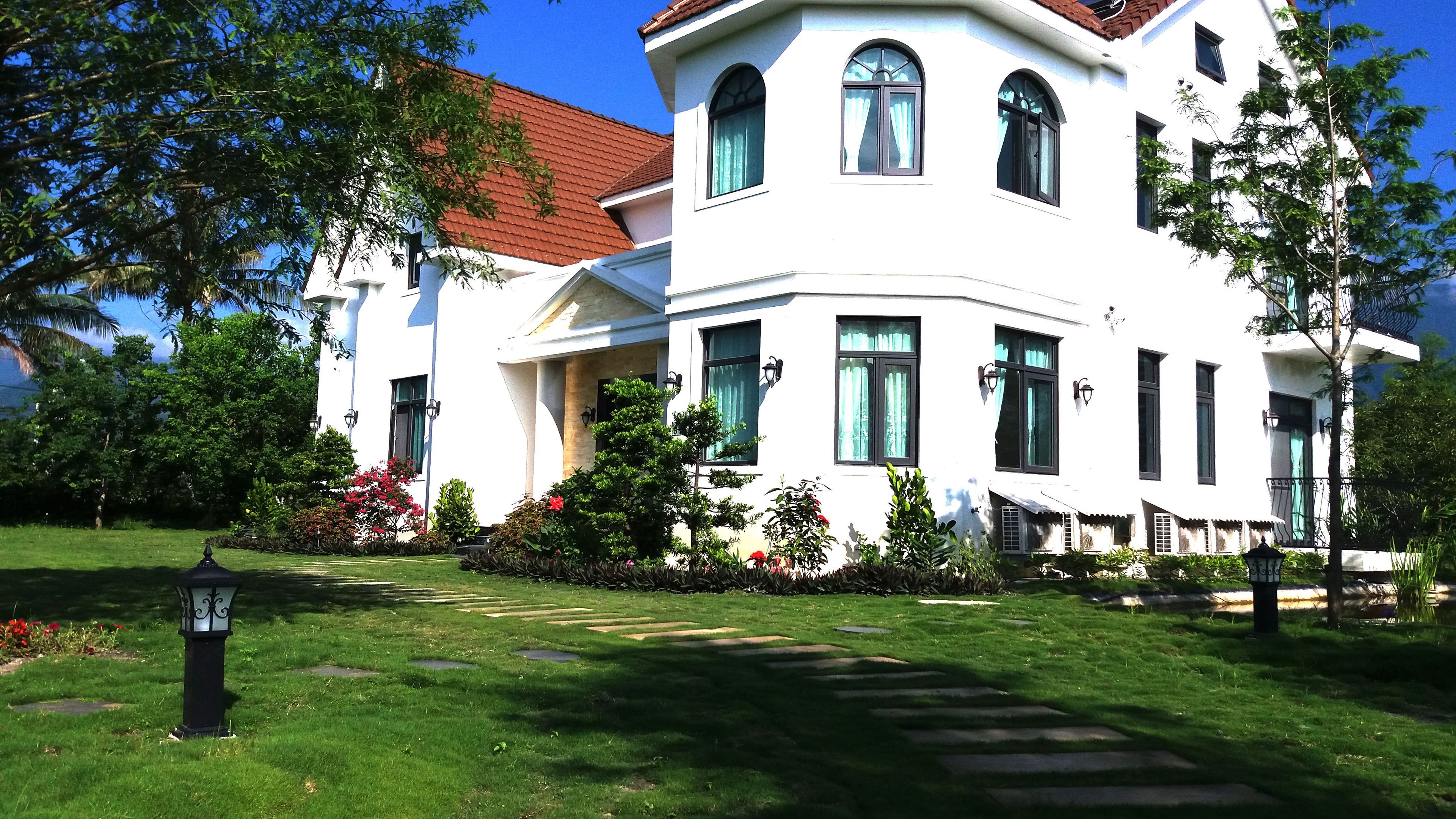 A two-story white building with a red-tiled roof, surrounded by a well-maintained garden with various plants and flowers.