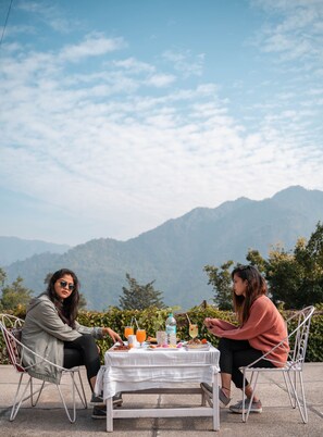 Two people seated at a table with a white tablecloth, surrounded by greenery and mountains in the background. - Swiss Cottage and Spa by Salvus (Narendranagar)