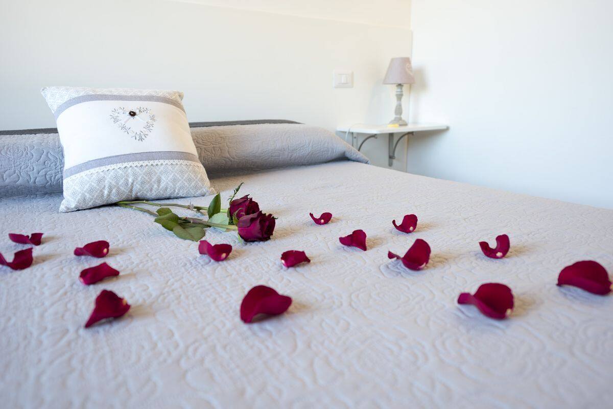 A neatly made bed with a white textured bedspread, a decorative pillow with a heart design, and scattered rose petals.