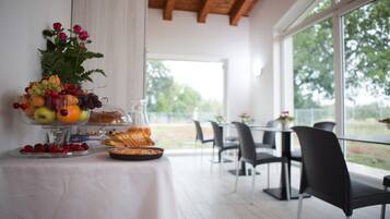 A breakfast buffet with fruits, pastries, and a flower arrangement on a table in a bright room with large windows.