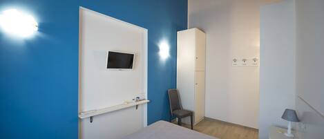 A bedroom with a bed, a chair, a TV mounted on the wall, a wardrobe, and a bedside table with a lamp.